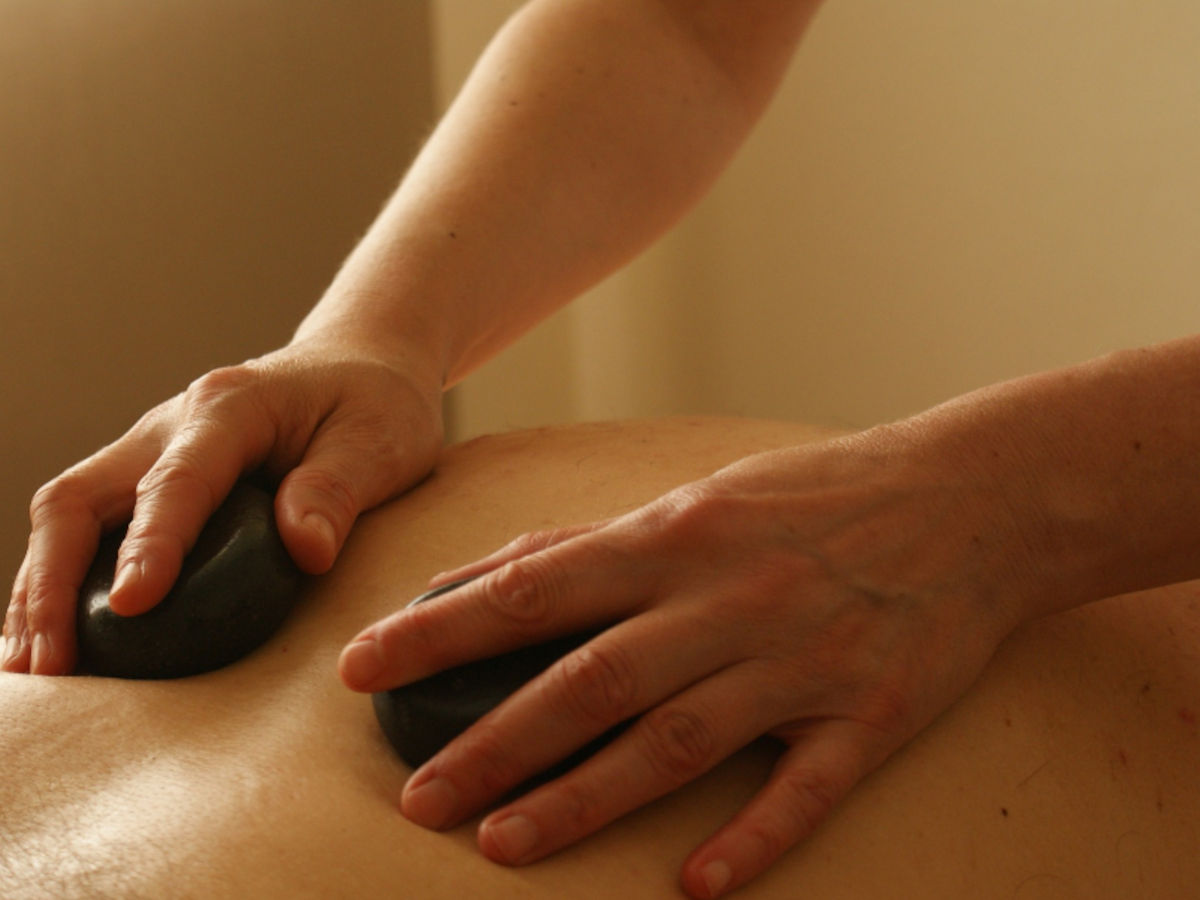 A person having hot stone massage by a therapist holding 2 black hot stones with both hands on the back.